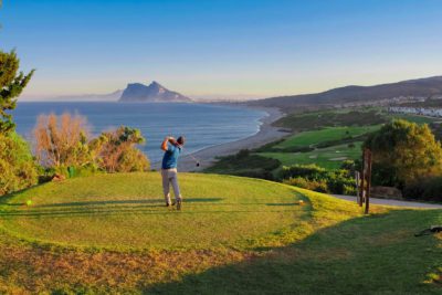 Person teeing off with trees around and a view of sandy beaches and mountains in the background