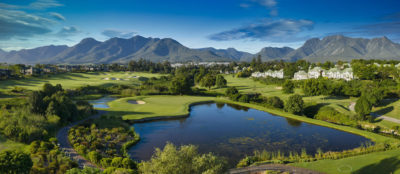 Aerial view of a lake on a fairway with trees around and buildings and mountains in the distance