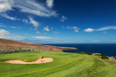 Person chipping out of a bunker on to a green with a white flag, overlooking the ocean