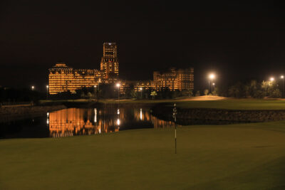 Hole with lake next to it at Al Hamra Golf Club with building in distance at night